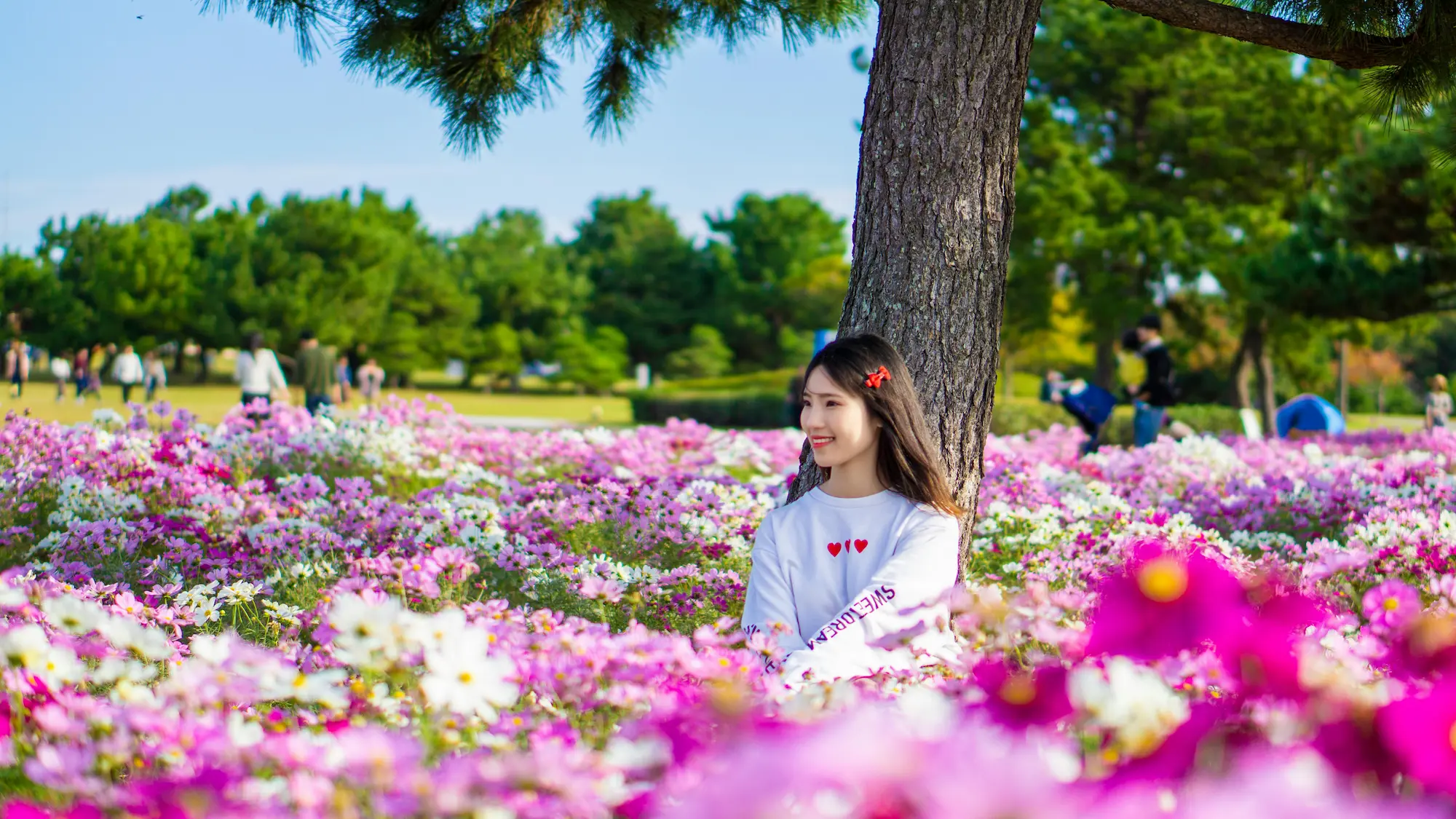 A young woman sits under a tree in a field of pink and white flowers, quietly reflecting on her self-awareness, with people and greenery visible in the background on a sunny day.